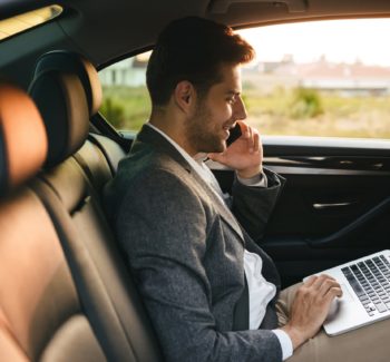 Confident businessman talking on mobile phone while sitting with laptop computer at the back of a car